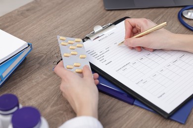 Photo of Doctor with pen and blister of pills filling out patient's medical card at wooden table, closeup