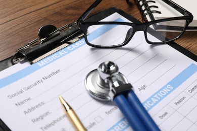 Photo of Medical card form, clipboard, pen and glasses on wooden table, closeup