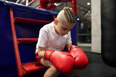 Sad boy in protective gloves sitting near boxing ring