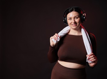 Photo of Plus size woman in gym clothes with headphones and towel on black background, space for text