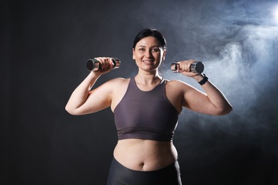 Photo of Plus size woman in gym clothes holding dumbbells on dark background with smoke