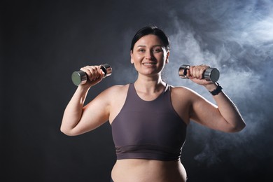 Photo of Plus size woman in gym clothes holding dumbbells on dark background with smoke