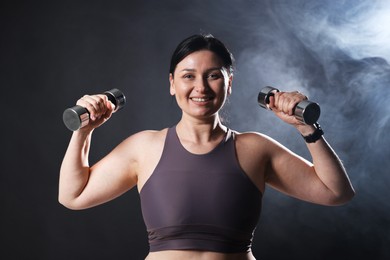 Photo of Plus size woman in gym clothes holding dumbbells on dark background with smoke