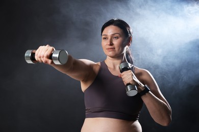 Photo of Plus size woman in gym clothes holding dumbbells on dark background with smoke