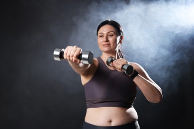 Photo of Plus size woman in gym clothes holding dumbbells on dark background with smoke