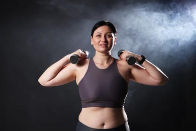 Photo of Plus size woman in gym clothes holding dumbbells on dark background with smoke