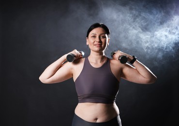 Photo of Plus size woman in gym clothes holding dumbbells on dark background with smoke