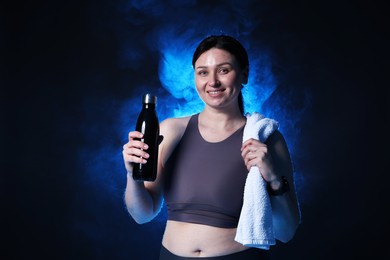 Photo of Plus size woman in gym clothes with water bottle and towel on dark blue background with smoke