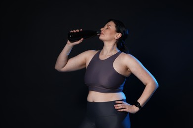 Photo of Plus size woman in gym clothes drinking water on black background