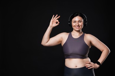 Photo of Plus size woman in gym clothes and headphones showing ok gesture on black background, space for text