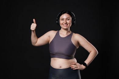 Photo of Plus size woman in gym clothes and headphones showing thumbs up on black background, space for text