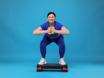 Photo of Plus size woman in gym clothes doing exercise with dumbbells and step platform on light blue background