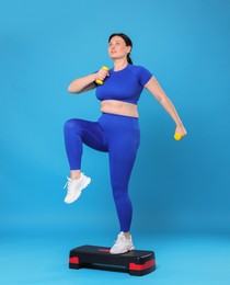 Photo of Plus size woman in gym clothes doing exercise with dumbbells and step platform on light blue background