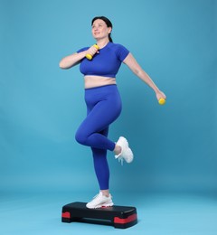 Photo of Plus size woman in gym clothes doing exercise with dumbbells and step platform on light blue background