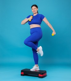 Photo of Plus size woman in gym clothes doing exercise with dumbbells and step platform on light blue background