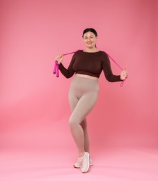 Plus size woman in gym clothes with skipping rope on pink background