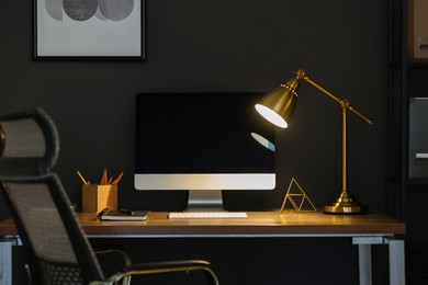 Photo of Comfortable workplace with computer and lamp on wooden desk at home office in evening