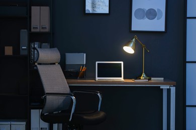 Photo of Comfortable workplace with laptop and lamp on wooden desk at home office in evening