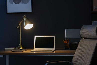 Photo of Comfortable workplace with laptop and lamp on wooden desk at home office in evening