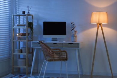 Photo of Comfortable workplace with computer on desk and floor lamp at home office in evening