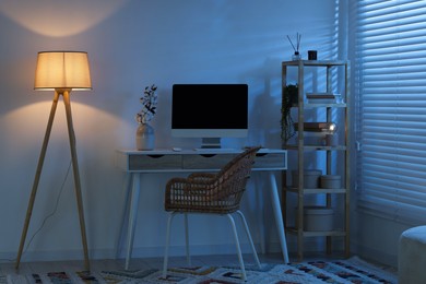 Photo of Comfortable workplace with computer on desk and floor lamp at home office in evening