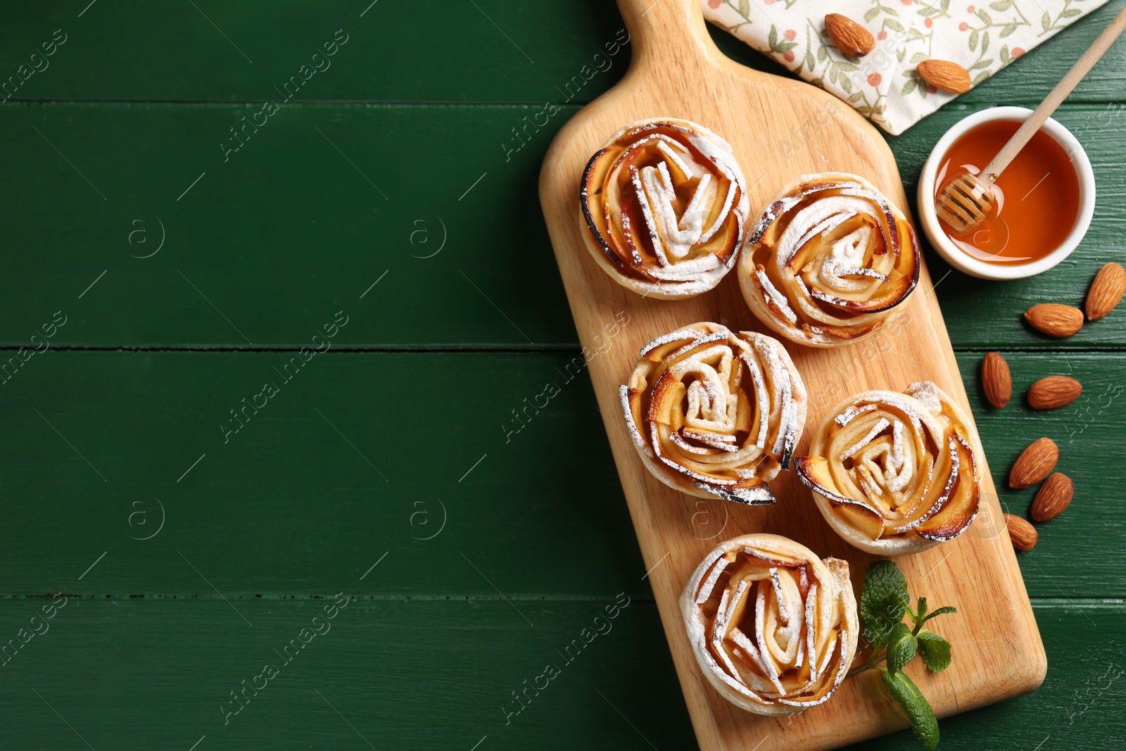 Photo of Tasty apple roses with powdered sugar served on green wooden table, flat lay. Space for text
