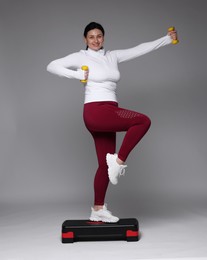 Photo of Plus size woman in gym clothes doing exercise with dumbbells and step platform on grey background