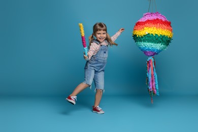 Happy girl with bright pinata and stick on light blue background