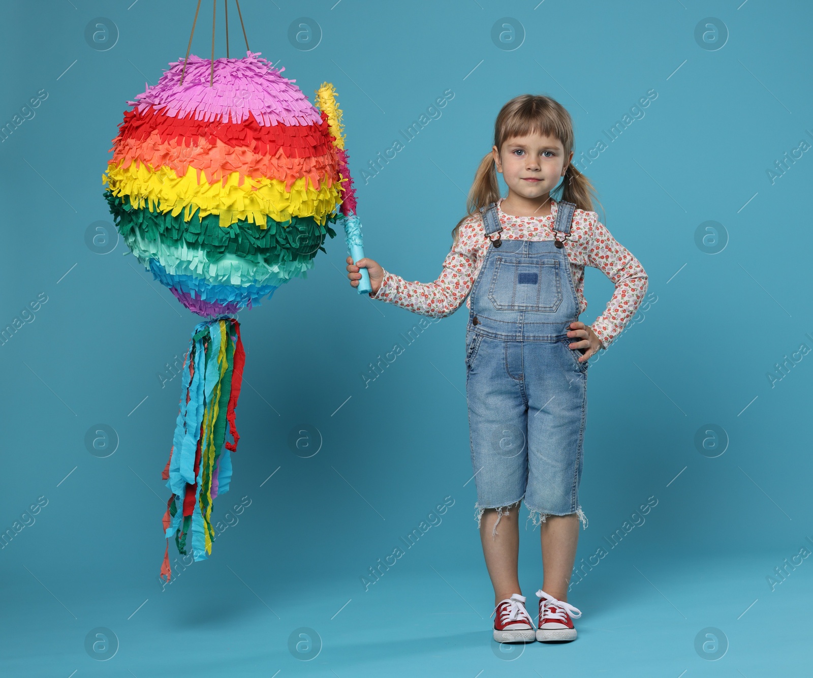 Photo of Cute girl with bright pinata and stick on light blue background