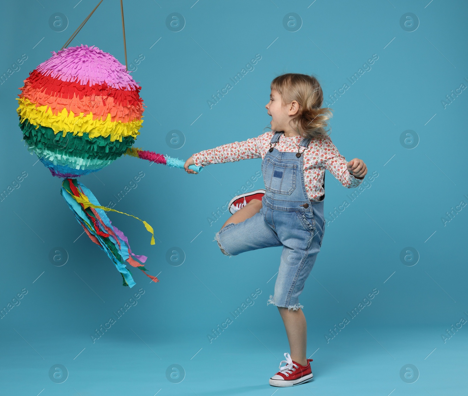 Photo of Happy girl breaking bright pinata with stick on light blue background