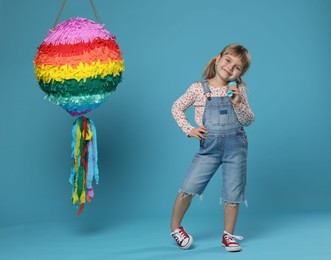 Photo of Cute girl with bright pinata and stick on light blue background