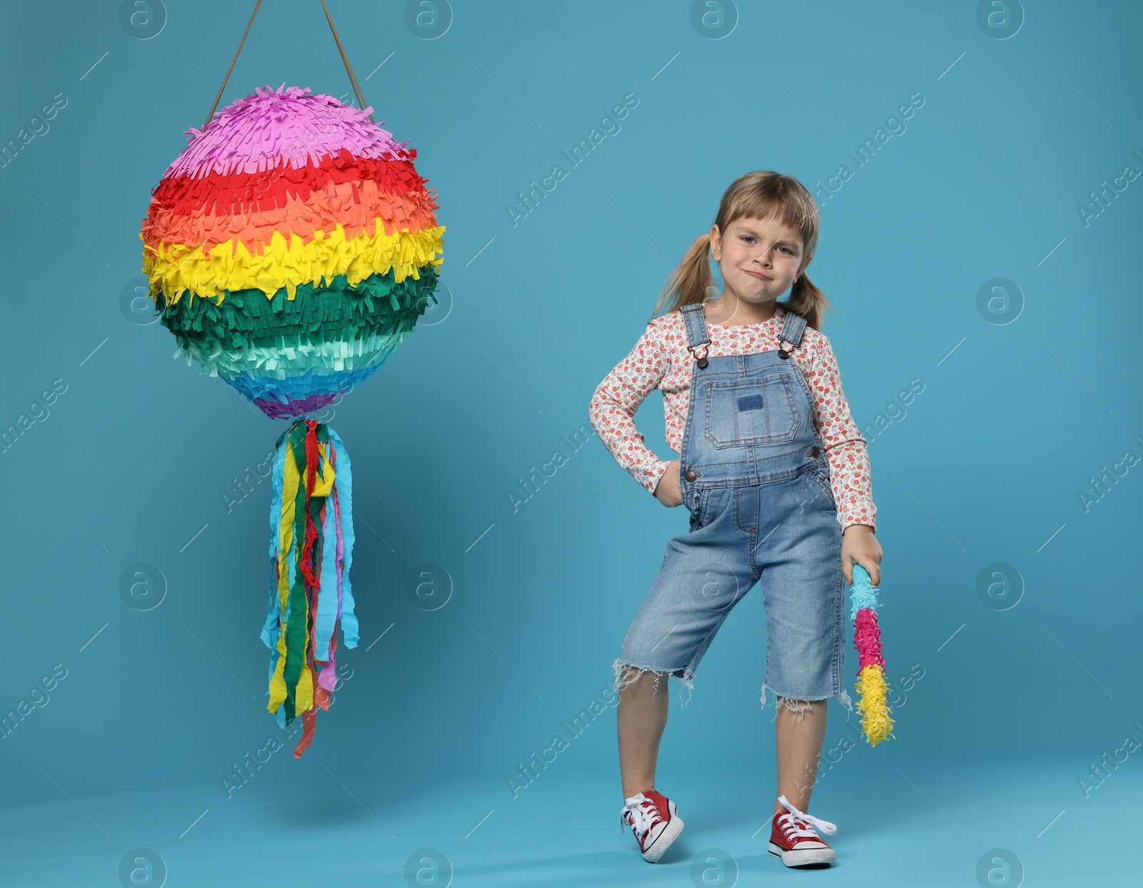 Photo of Cute girl with bright pinata and stick on light blue background