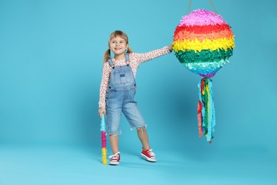 Happy girl with bright pinata and stick on light blue background