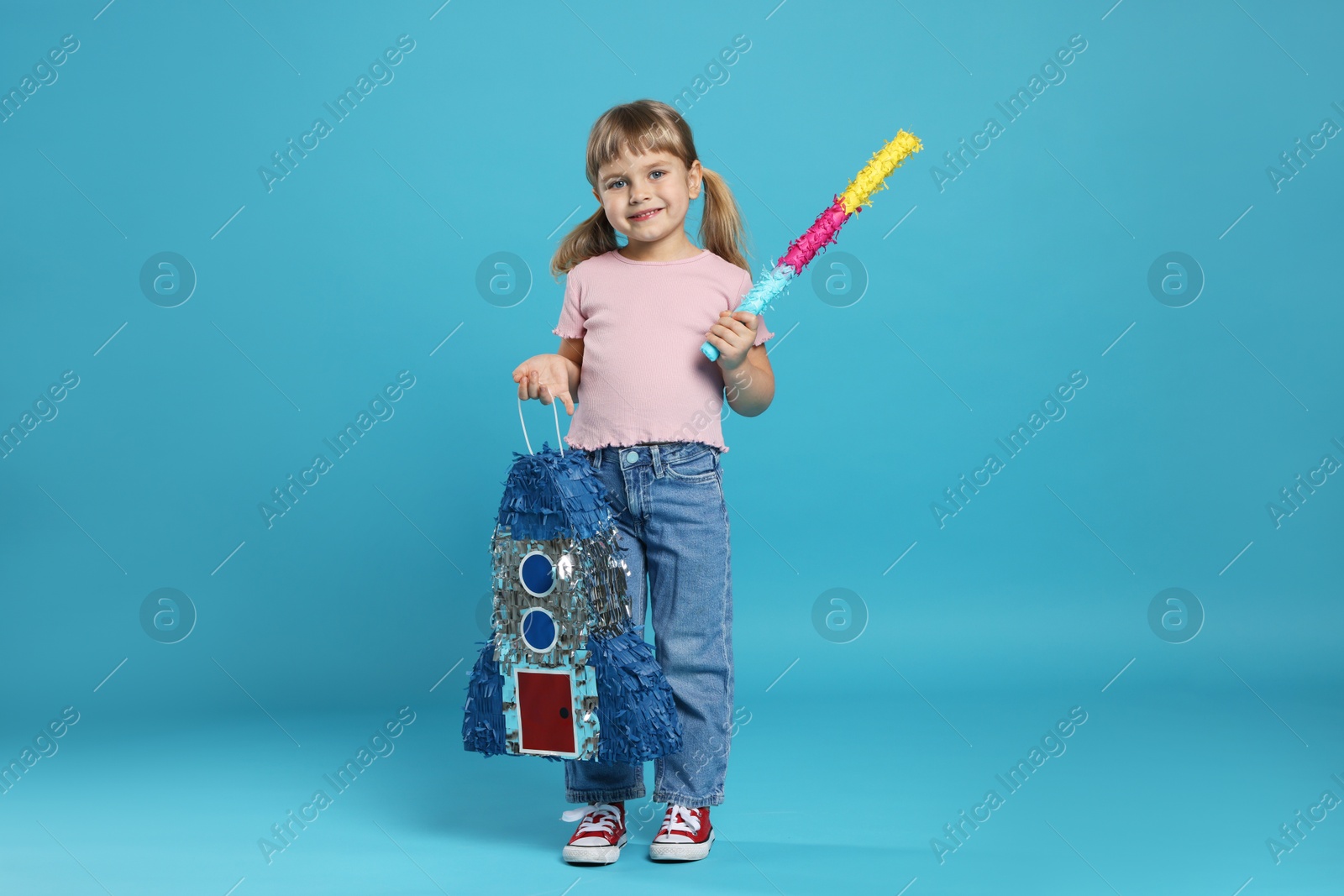 Photo of Happy girl with rocket shaped pinata and stick on light blue background