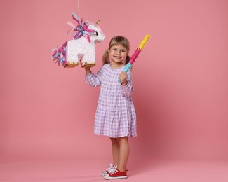 Happy girl with unicorn shaped pinata and stick on pink background