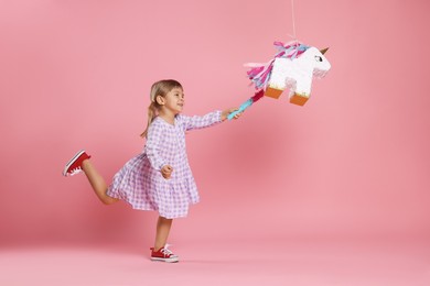 Happy girl breaking unicorn shaped pinata with stick on pink background