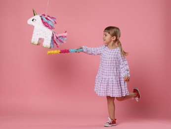 Happy girl breaking unicorn shaped pinata with stick on pink background