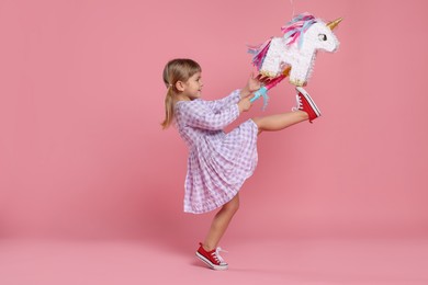 Cute girl breaking unicorn shaped pinata with stick on pink background