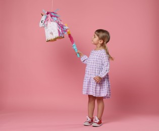 Cute girl with unicorn shaped pinata and stick on pink background