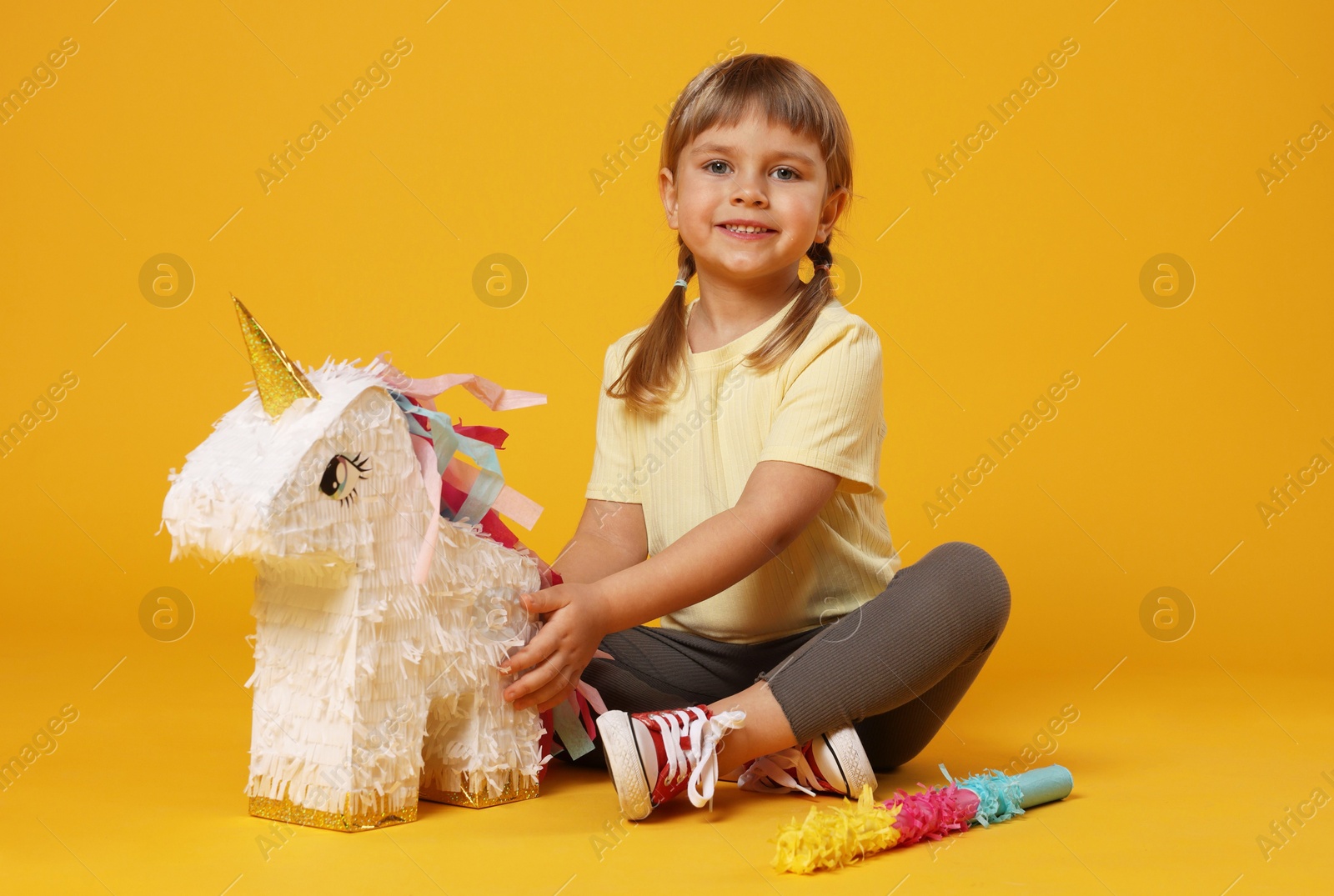 Photo of Happy girl with unicorn shaped pinata and stick on orange background