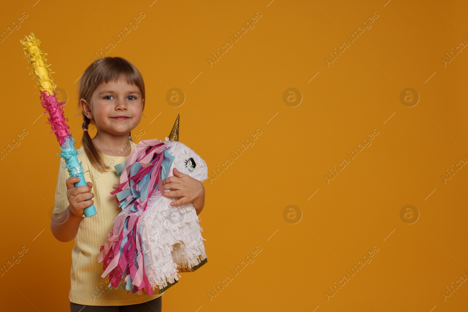 Photo of Happy girl with unicorn shaped pinata and stick on orange background. Space for text