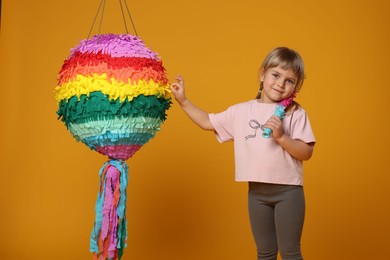 Photo of Cute girl with bright pinata and stick on orange background