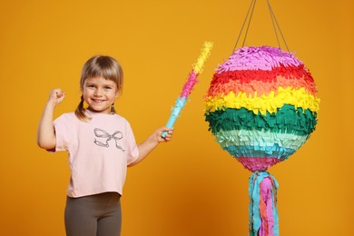 Happy girl with bright pinata and stick on orange background