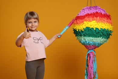 Photo of Cute girl with bright pinata and stick showing thumbs up on orange background