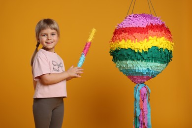 Happy girl with bright pinata and stick on orange background