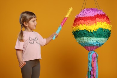 Happy girl with bright pinata and stick on orange background