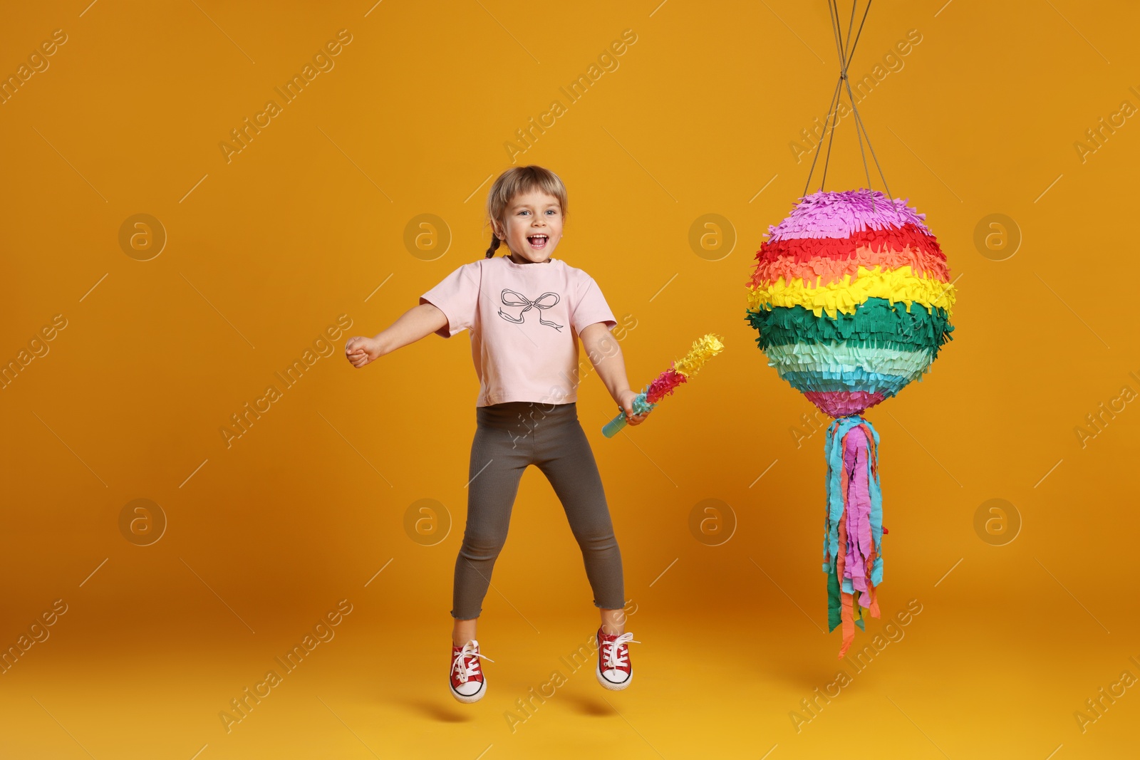 Photo of Happy girl with bright pinata and stick jumping on orange background