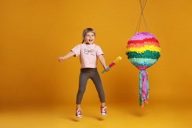 Happy girl with bright pinata and stick jumping on orange background