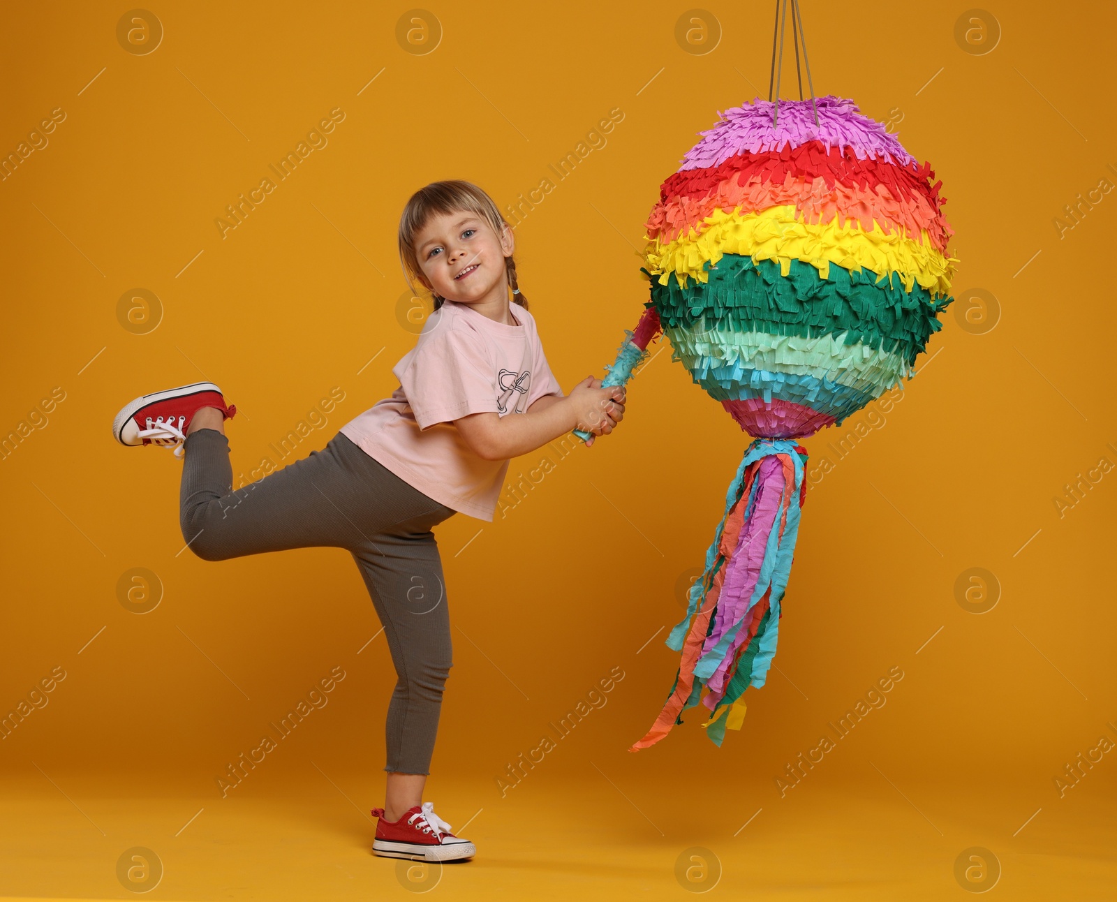 Photo of Cute girl breaking bright pinata with stick on orange background