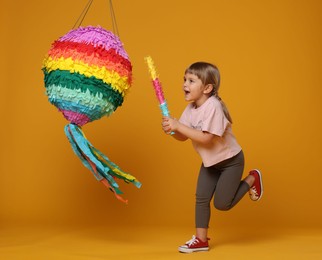 Photo of Cute girl breaking bright pinata with stick on orange background